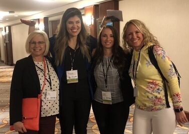 Four women standing indoors, smiling at the camera.