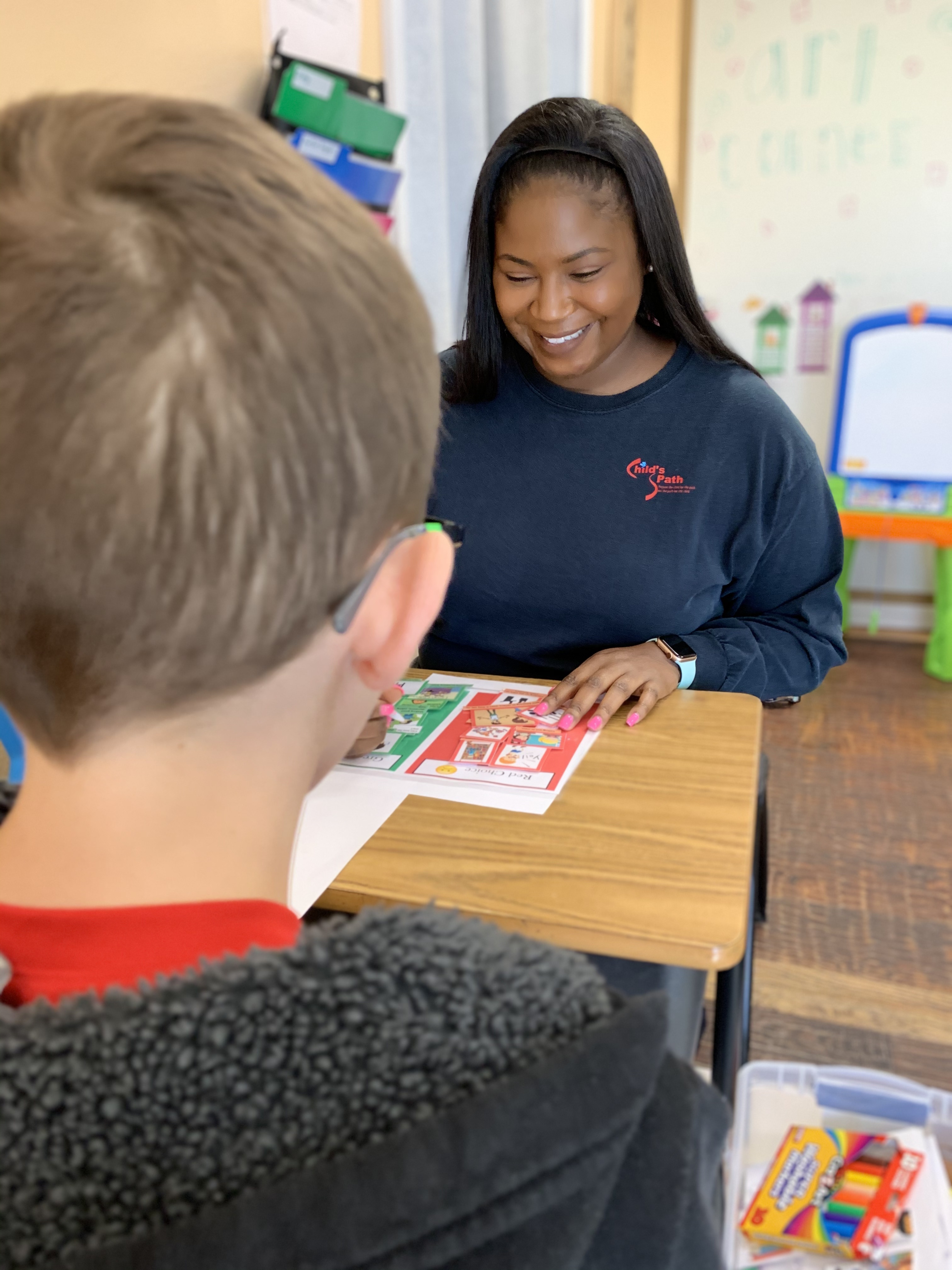 A teacher smiling while helping a student with a colorful worksheet in a classroom.