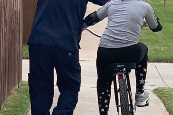 An adult helping a child ride a bicycle on a sidewalk.