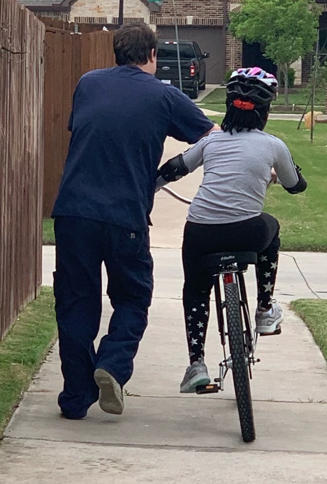 An adult helping a child ride a bicycle on a sidewalk.