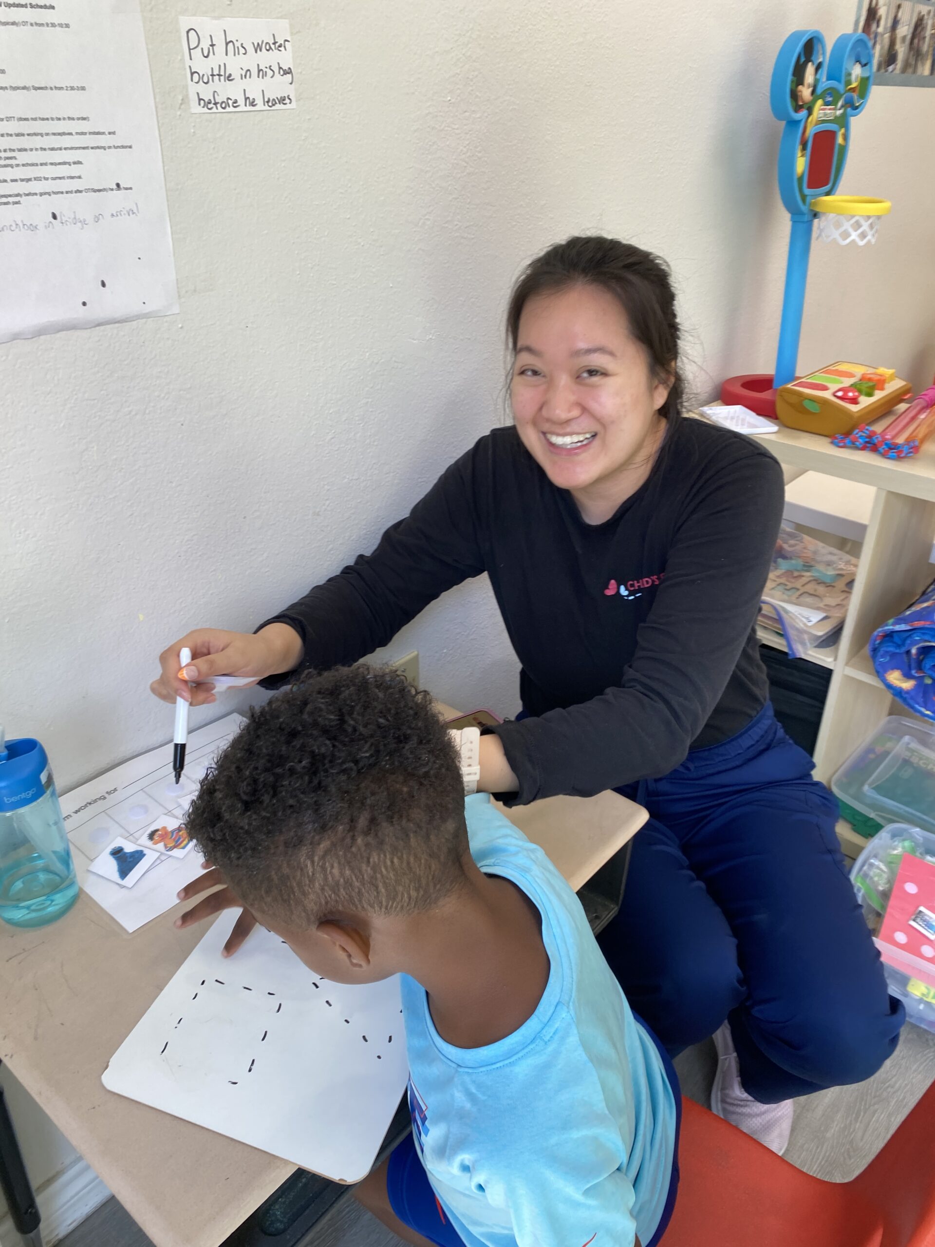 A teacher happily helps a young boy with a drawing activity at a small desk.