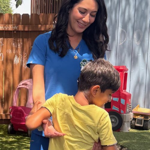 A woman in blue helps a young boy with a toy outdoors.