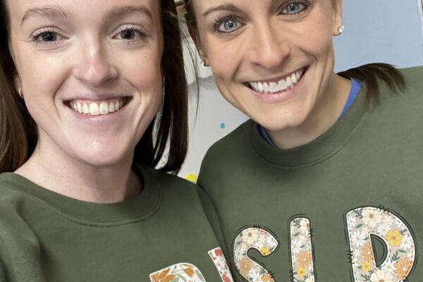 Two smiling women wearing matching green shirts with floral text.